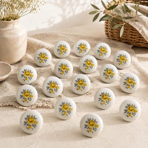 May include: A collection of sixteen round white ceramic cabinet knobs, each featuring a central yellow rose motif surrounded by small grey leaves. The knobs are arranged in a grid pattern, displaying a consistent floral design on their smooth surfaces.