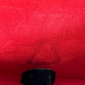 Puede incluir: Un anillo negro con una cara rectangular se muestra sobre un fondo rojo vibrante. El anillo parece estar hecho de un material oscuro, posiblemente similar al terciopelo. El marco es una caja roja forrada de tela.