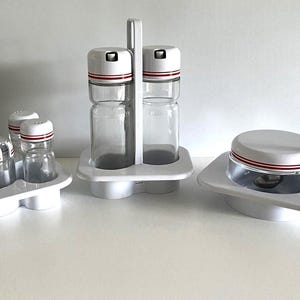 May include: A set of clear glass kitchen canisters with white lids and red stripes. The set includes a double canister with a handle, a single canister with a spoon, and a double canister set. All are in white trays.