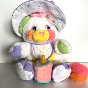 May include: A vintage plush toy bear with a sun hat, bib, bottle, and a small round object. The bear is white with colorful accents of pink, orange, yellow, green, and purple. The hat and bib have a star pattern.