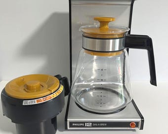 May include: A Philips 12 Dial-A-Brew coffee maker with a silver and black finish, featuring a glass carafe with a yellow lid and a separate black filter basket component with a yellow dial and orange markings for brew strength settings.