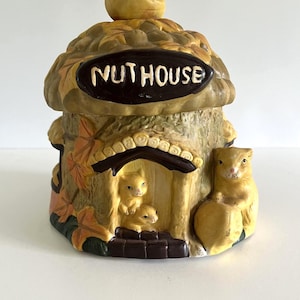 Peut inclure: Un pot en céramique en forme de gland avec un couvercle surmonté d'une noix. Le pot porte l'inscription "NUTHOUSE" sur une plaque marron foncé, avec des écureuils sculptés dans une porte et sur le côté, ornés de feuilles d'automne peintes.