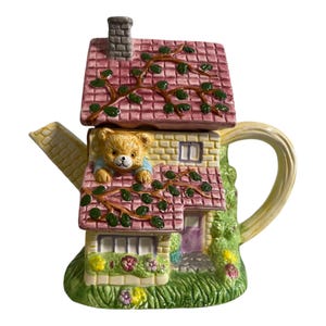 May include: A ceramic teapot shaped like a cottage with a pink tiled roof, a grey chimney, and green vines. A brown teddy bear peers out from an upper window. The base features yellow and purple flowers, a purple door, and a yellow spout and handle.