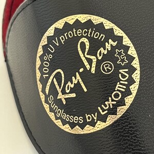 Puede incluir: Funda negra para gafas de sol con un emblema dorado que dice "Ray-Ban Sunglasses by Luxottica" y "100% UV protection". La funda tiene un interior rojo.