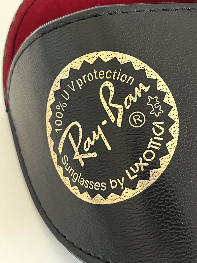 Puede incluir: Funda negra para gafas de sol con un emblema dorado que dice "Ray-Ban Sunglasses by Luxottica" y "100% UV protection". La funda tiene un interior rojo.