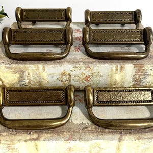 May include: A set of six brass-toned metal drawer pulls featuring a rectangular central plate with a textured finish, bordered by a beaded edge, and a curved drop handle. Each pull has a polished metallic surface with a warm, aged appearance.