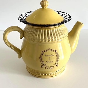 May include: Yellow ceramic teapot with a black wire rim and lid. The teapot has a curved handle and spout. The front of the teapot features the text "Pasticceria di Venezia" within a decorative floral wreath.