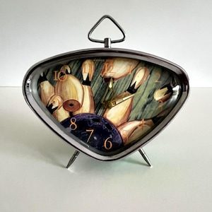 May include: A retro-style alarm clock with a silver metal frame and a unique bowling-themed face. The clock face features a bowling ball, pins, and a green and brown background. The clock has a triangular top handle and three small legs.