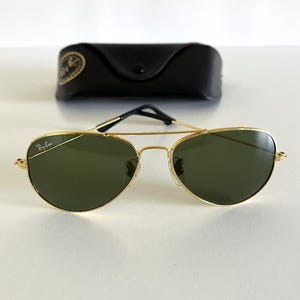 Puede incluir: Gafas de sol aviador con montura dorada, lentes verdes y estuche negro. Las gafas tienen doble puente y el logotipo de Ray-Ban en la lente y la montura. El estuche es negro con un emblema dorado.