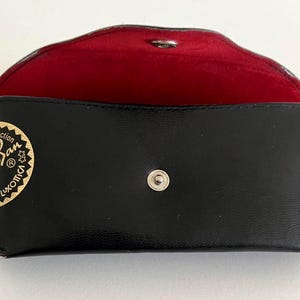 Puede incluir: Estuche rectangular negro para gafas de sol con interior rojo. El estuche tiene un cierre a presi&oacute;n y un logotipo dorado que dice "Ray-Ban" y "Luxottica". El estuche est&aacute; abierto, revelando el forro rojo.