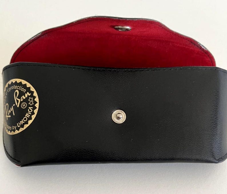 Puede incluir: Estuche rectangular negro para gafas de sol con interior rojo. El estuche tiene un cierre a presi&oacute;n y un logotipo dorado que dice "Ray-Ban" y "Luxottica". El estuche est&aacute; abierto, revelando el forro rojo.
