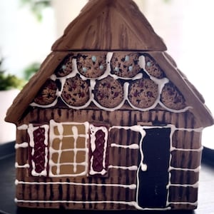 Peut inclure: Un pot à biscuits en céramique marron en forme de maison en pain d'épices. Le toit est marron avec des accents de biscuits. La maison a des détails de glaçage blanc, une porte noire et des fenêtres avec des accents rouges et jaunes. Il repose sur un plateau noir.