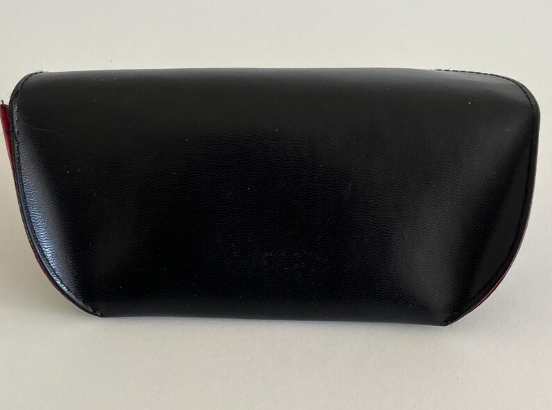 Puede incluir: Un estuche para gafas negro, de aspecto cuero, con forma curvada. El estuche tiene un acabado liso y brillante y un acento rojo en el lateral. El estuche est&aacute; cerrado y parece estar vac&iacute;o.