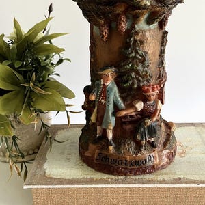 Peut inclure: Un récipient décoratif cylindrique en résine brune présentant une sculpture en relief d'une personne en manteau bleu et d'une personne en tenue rouge et noire près d'un sapin. La base porte le mot Schwarzwald en relief sur une bannière.