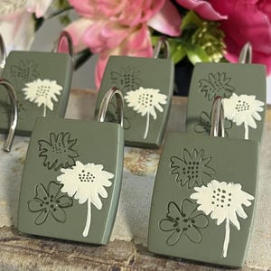 Peut inclure: Un ensemble de crochets de rideau de douche vert sauge, chacun présentant une face rectangulaire avec un motif floral en creux. Une fleur est mise en évidence en blanc, tandis que deux fleurs plus petites sont gravées sur la surface verte.