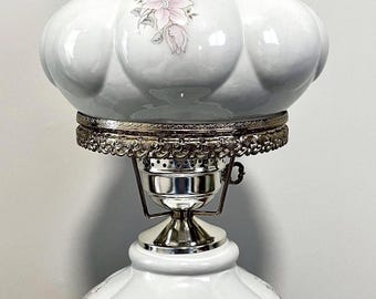 May include: A white ceramic hurricane lamp featuring a scalloped glass shade and a matching base, both decorated with delicate pink floral motifs. The lamp includes a silver-toned metal collar with intricate filigree detailing and a central socket.