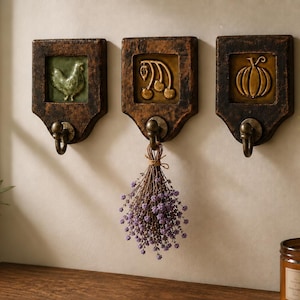May include: Three dark brown wooden wall hooks with square decorative tiles. The left tile features a green chicken, the center a gold cherry cluster, and the right a gold pumpkin. Each hook is made of aged wood with a metal hook attached at the base.