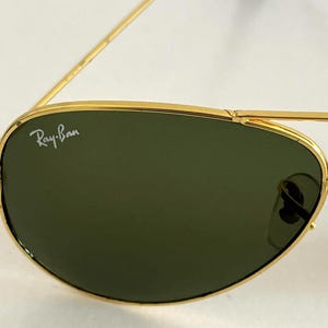 Puede incluir: Primer plano de gafas de sol Ray-Ban con montura dorada y lentes verdes. El logotipo de Ray-Ban es visible en la lente izquierda. Las gafas de sol tienen un estilo aviador cl&aacute;sico.