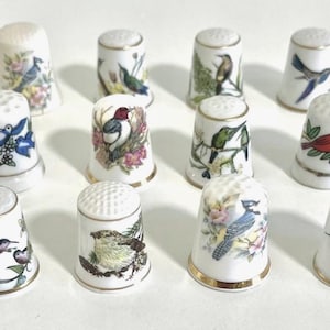 May include: A set of twelve white porcelain thimbles, each decorated with a different colourful bird illustration. The thimbles feature gold rims and detailed bird designs, showcasing various species and floral accents.