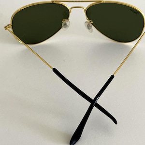 Puede incluir: Gafas de sol de aviador con montura dorada y lentes verde oscuro. Las gafas tienen un doble puente y patillas negras. El logotipo de Ray-Ban es visible en la lente derecha. Sobre fondo blanco.