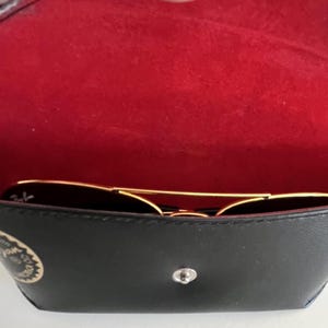 Puede incluir: Gafas de sol negras con montura dorada dentro de un estuche negro abierto con interior rojo. El estuche, con cierre plateado y logotipo, es parcialmente visible. Las gafas son de estilo aviador cl&aacute;sico.