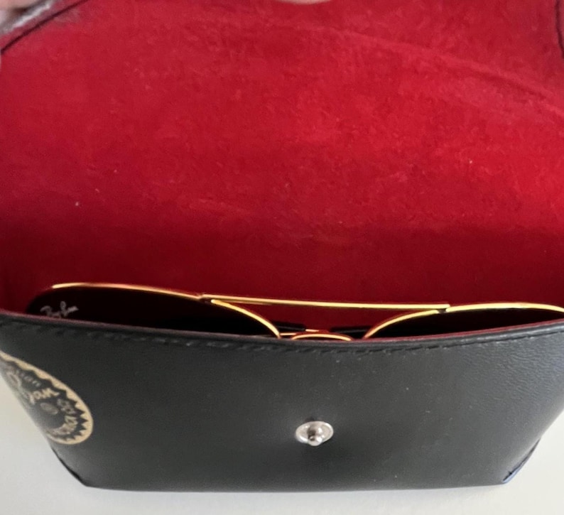 Puede incluir: Gafas de sol negras con montura dorada dentro de un estuche negro abierto con interior rojo. El estuche, con cierre plateado y logotipo, es parcialmente visible. Las gafas son de estilo aviador cl&aacute;sico.