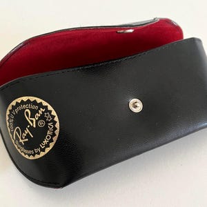 Puede incluir: Estuche negro para gafas de sol con interior rojo. El estuche est&aacute; abierto, revelando el forro rojo. El logotipo de Ray-Ban se muestra en dorado en la parte delantera. El estuche tiene un cierre a presi&oacute;n.