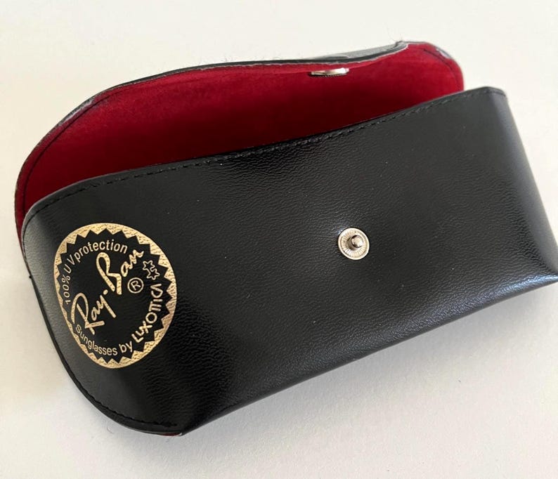 Puede incluir: Estuche negro para gafas de sol con interior rojo. El estuche est&aacute; abierto, revelando el forro rojo. El logotipo de Ray-Ban se muestra en dorado en la parte delantera. El estuche tiene un cierre a presi&oacute;n.