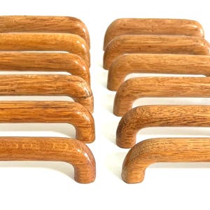 Peut inclure: Un ensemble de douze poignées de placard en bois assorties, disposées en deux rangées parallèles. Chaque poignée présente un design arqué et lisse avec une finition en grain de bois brun clair naturel, affichant une texture et une forme courbée.