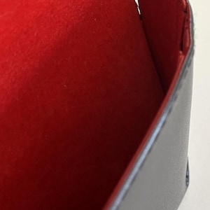 Puede incluir: Un estuche rectangular negro con un interior rojo vibrante. El estuche parece estar hecho de cuero o un material similar, con costuras visibles en los bordes. El interior es un material suave, similar al fieltro.
