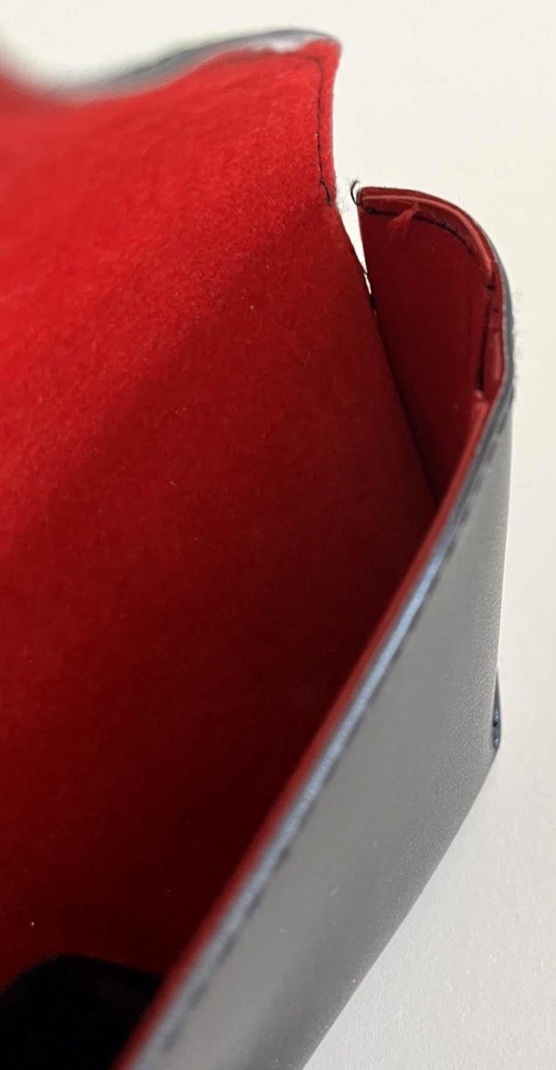 Puede incluir: Un estuche rectangular negro con un interior rojo vibrante. El estuche parece estar hecho de cuero o un material similar, con costuras visibles en los bordes. El interior es un material suave, similar al fieltro.