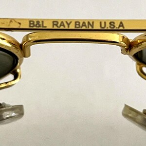 Puede incluir: Primer plano de gafas de sol con montura dorada y lentes oscuras. La barra superior de la montura lleva la inscripci&oacute;n "B&L RAY BAN U.S.A.". Las gafas de sol tienen un dise&ntilde;o cl&aacute;sico, con puente y varillas doradas.