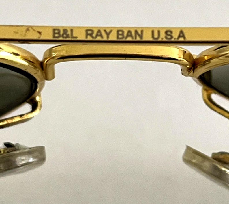 Puede incluir: Primer plano de gafas de sol con montura dorada y lentes oscuras. La barra superior de la montura lleva la inscripci&oacute;n "B&L RAY BAN U.S.A.". Las gafas de sol tienen un dise&ntilde;o cl&aacute;sico, con puente y varillas doradas.