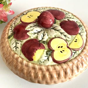 Peut inclure: Plat à tarte en céramique avec un bord en forme de panier tressé. La tarte est décorée de tranches de pommes rouges et jaunes et de pommes entières. Des feuilles vertes sont parsemées parmi les fruits. Un article de cuisine décoratif.