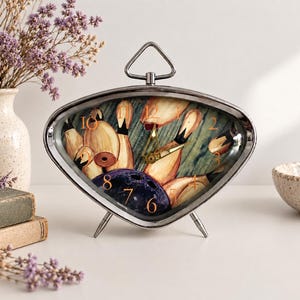May include: A silver-toned, triangular-shaped analog table clock with a metallic frame and a top handle. The clock face features a graphic print of bowling pins and a dark blue bowling ball, with orange numerals and gold-colored clock hands.