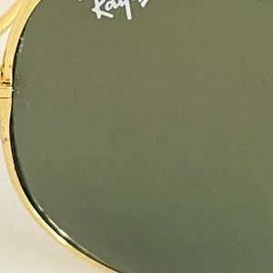 Puede incluir: Primer plano de gafas de sol Ray-Ban. Las gafas de sol tienen una montura dorada y lentes verdes. El logotipo de Ray-Ban es visible en blanco en la lente superior izquierda. La imagen est&aacute; bien iluminada, destacando los detalles de las gafas.