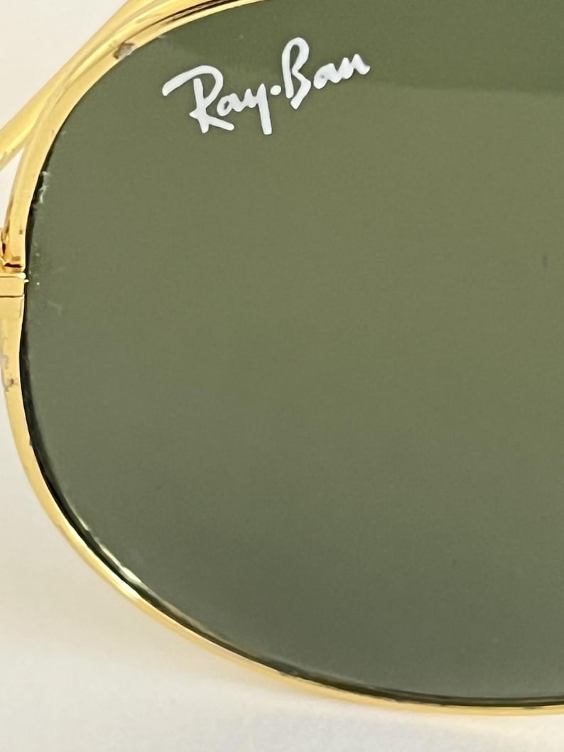 Puede incluir: Primer plano de gafas de sol Ray-Ban. Las gafas de sol tienen una montura dorada y lentes verdes. El logotipo de Ray-Ban es visible en blanco en la lente superior izquierda. La imagen est&aacute; bien iluminada, destacando los detalles de las gafas.