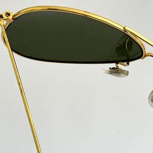 Puede incluir: Primer plano de gafas de sol con montura dorada y lentes verde oscuro. Las gafas de sol tienen una forma de aviador cl&aacute;sica, con una montura dorada fina y un puente. Las lentes son de color verde oscuro.