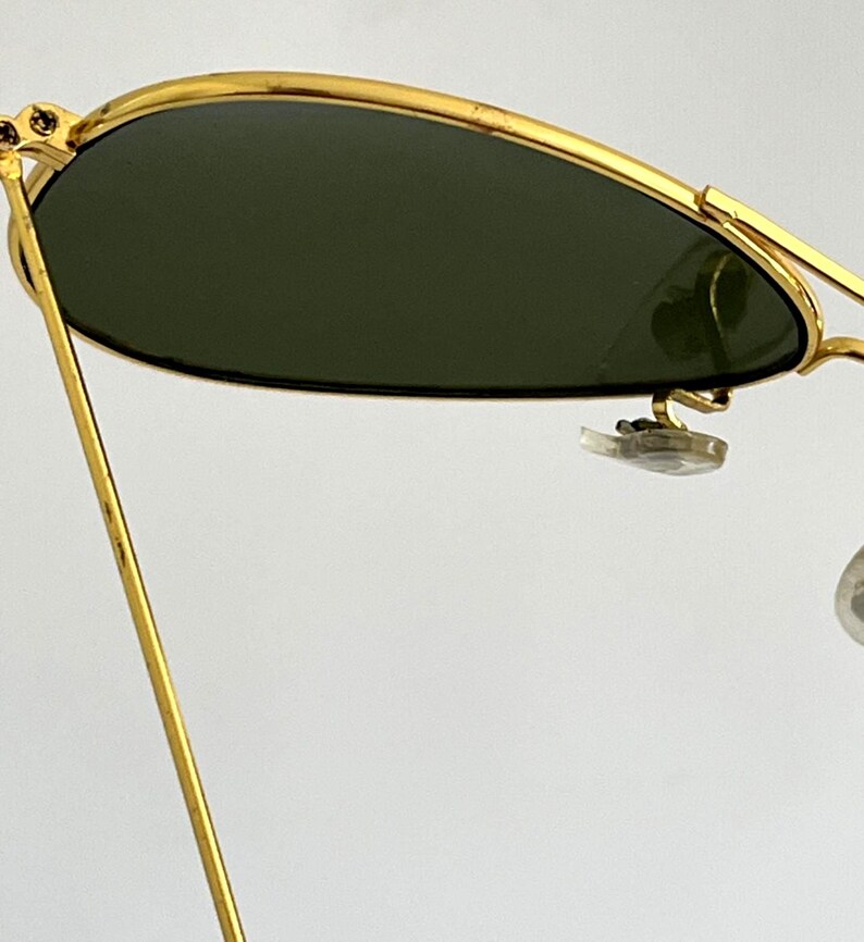 Puede incluir: Primer plano de gafas de sol con montura dorada y lentes verde oscuro. Las gafas de sol tienen una forma de aviador cl&aacute;sica, con una montura dorada fina y un puente. Las lentes son de color verde oscuro.