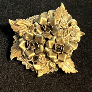 Peut inclure: Une broche en métal doré représentant trois fleurs de rose en relief et des feuilles détaillées superposées sur une base carrée, géométrique et ajourée. La surface métallique présente une finition brunie avec des reflets.