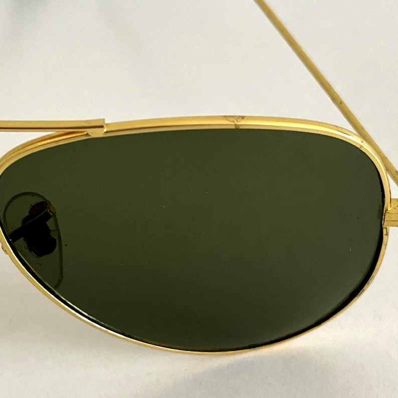 Puede incluir: Primer plano de gafas de sol con montura dorada y lentes verdes. Las gafas de sol tienen un dise&ntilde;o cl&aacute;sico de aviador, con doble puente y finas varillas doradas. Las lentes son de color verde oscuro.