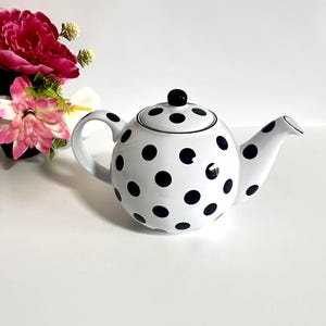 Peut inclure: Théière en céramique blanche à pois noirs. La théière a un corps arrondi, un bec verseur incurvé et une anse en boucle. Un bouton noir est posé sur le couvercle. La théière est posée sur un fond blanc, avec des fleurs roses et rouges en arrière-plan.