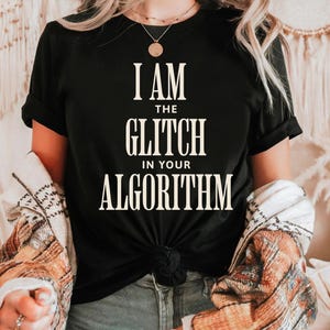 Pode incluir: T-shirt preto com o texto branco "I AM THE GLITCH IN YOUR ALGORITHM". A camisa está amarrada na cintura. A pessoa está usando uma jaqueta estampada e jeans.