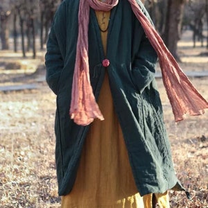 May include: A dark green quilted jacket with a long, flowing pink scarf. The jacket has a button closure and pockets on the sides. The person is wearing a yellow dress underneath the jacket.