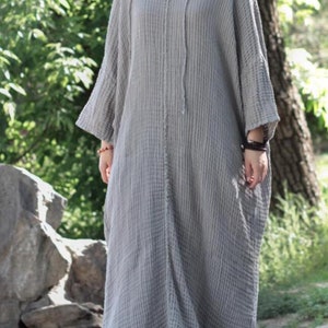 May include: A gray, long-sleeved, loose-fitting dress with a V-neck and a drawstring hood. The dress is made of a textured fabric and has a relaxed fit.