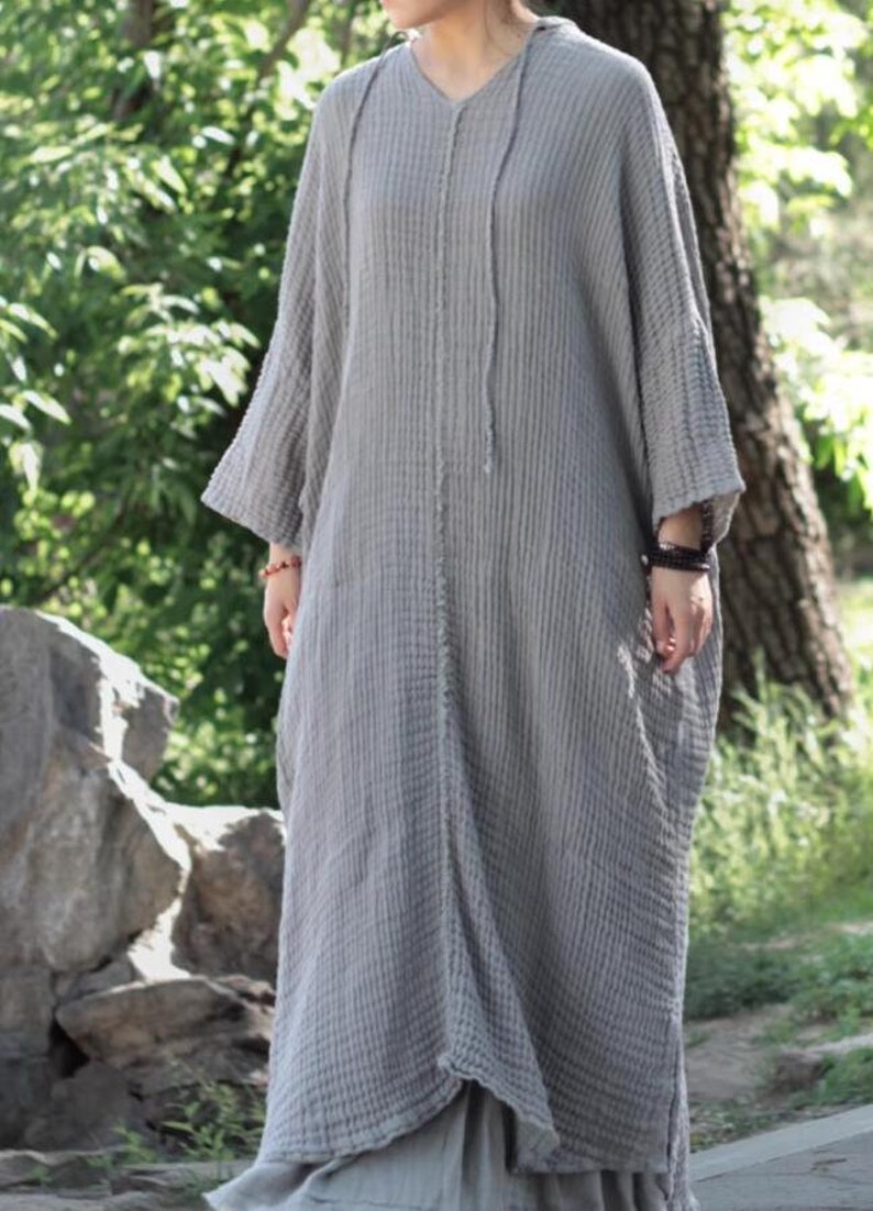 May include: A gray, long-sleeved, loose-fitting dress with a V-neck and a drawstring hood. The dress is made of a textured fabric and has a relaxed fit.