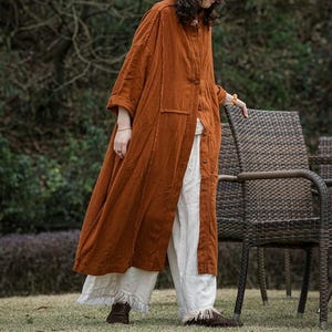 Op de afbeelding: Een lange, roestkleurige linnen jas met een knoopsluiting en een relaxte pasvorm. De jas heeft een opgestikte zak en wordt gecombineerd met een witte, wijde broek met gefranjerde zomen. De outfit wordt gecompleteerd met bruine schoenen.