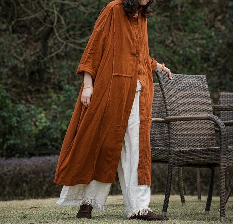 Op de afbeelding: Een lange, roestkleurige linnen jas met een knoopsluiting en een relaxte pasvorm. De jas heeft een opgestikte zak en wordt gecombineerd met een witte, wijde broek met gefranjerde zomen. De outfit wordt gecompleteerd met bruine schoenen.