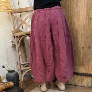 May include: Wide-leg, dusty rose-colored pants with a gathered waist and vertical seam details. The pants have a voluminous, balloon-like shape. The fabric appears to be a lightweight linen or cotton blend. The pants are paired with neutral-toned shoes.