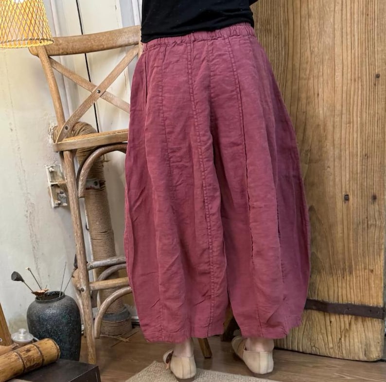 May include: Wide-leg, dusty rose-colored pants with a gathered waist and vertical seam details. The pants have a voluminous, balloon-like shape. The fabric appears to be a lightweight linen or cotton blend. The pants are paired with neutral-toned shoes.